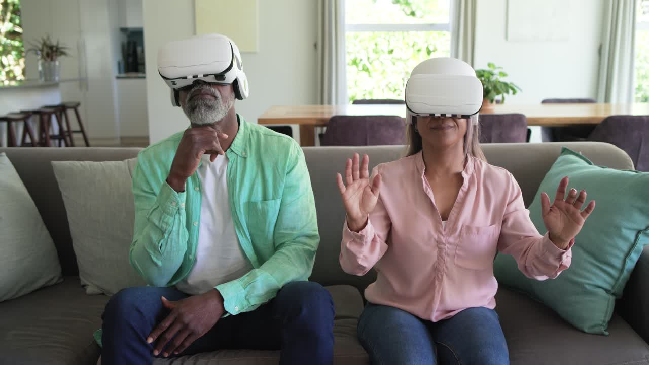 Coppia multietnica che indossa visori VR bianchi, raggiungendo ed esplorando oggetti virtuali sul divano