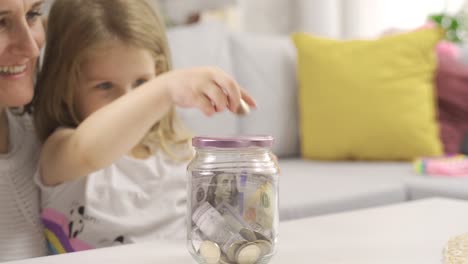 Little-girl-puts-money-in-piggy-bank-with-her-mother.-Her-mother-teaches-her-daughter-how-to-save-money.