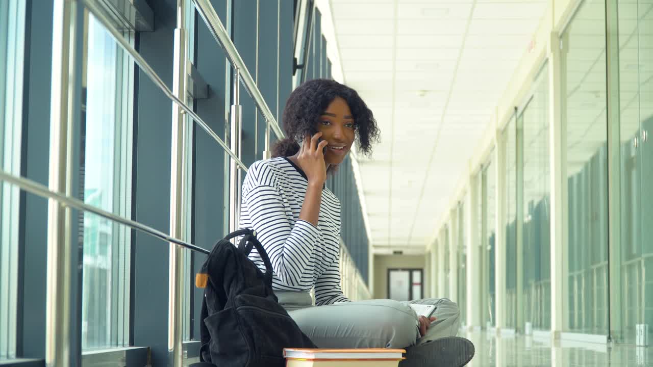 African american woman student sitting on the floor talking on the phone in the university coridor. New modern fully functional education facility. Concept of online education