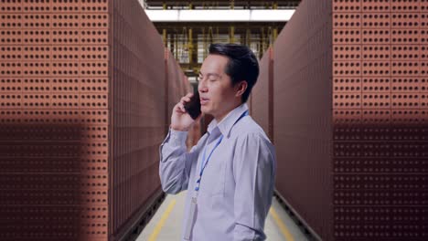 side view asian male professional worker standing with red brick packed in stacks are stored, he pick the phone up and started to specking to it with a slicely smile