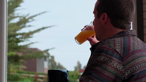 young-man-drinking-juice,-having-breakfast-on-cloudy-day,-forest-in-background,-hotel-breakfast,-airbnb,-Adult-male-enjoying-useful-drink-for-breakfast