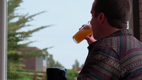 joven bebiendo jugo, desayunando en un día nublado, bosque al fondo, desayuno en el hotel, airbnb, hombre adulto disfrutando de una bebida útil para el desayuno