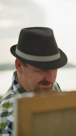close-up of a painter wearing a black hat, checkered shirt, and jeans, deeply focused on painting a large canvas set up in a tranquil grass field by a serene lake