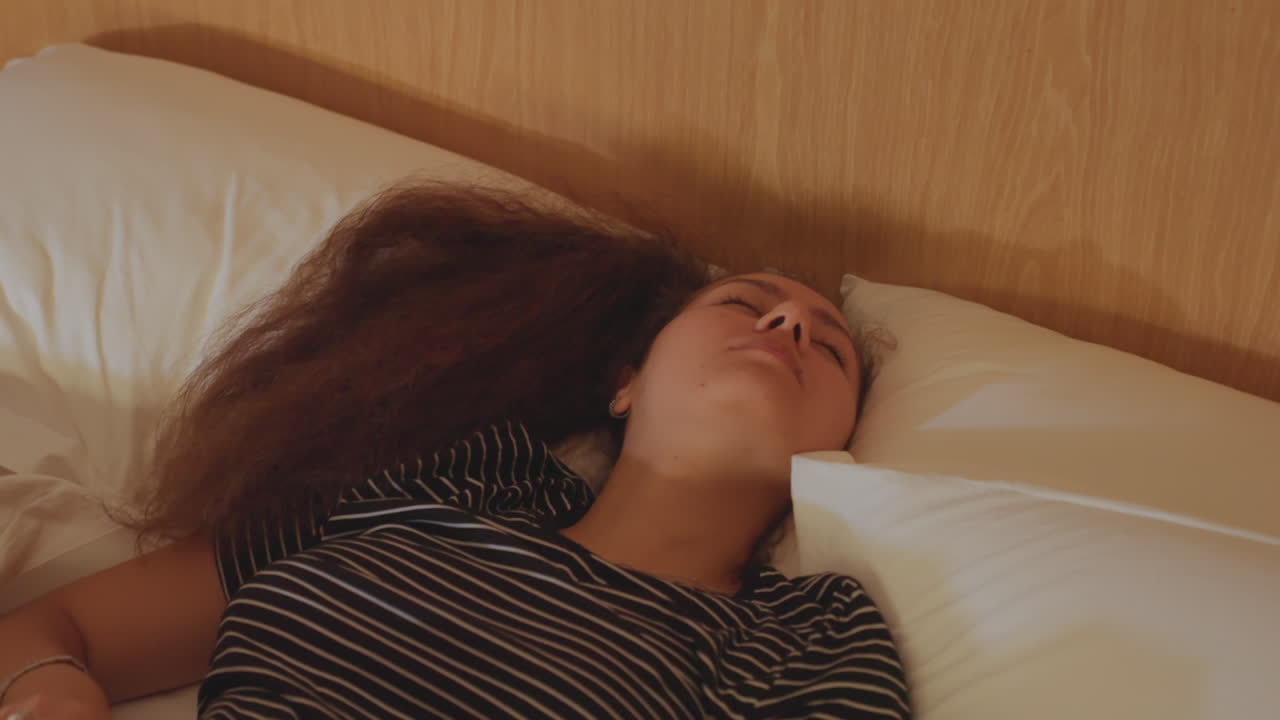 Guest sits thoughtfully on bed in hotel room. Calm woman with striped shirt relaxes in cozy hotel space. Quiet morning ambiance with reflective guest sitting on soft bed in hotel room