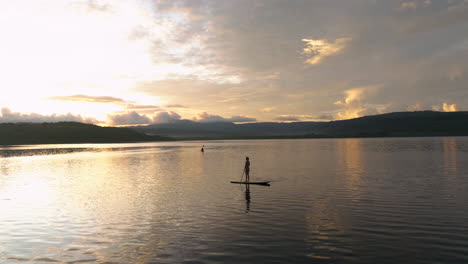 Mujer-De-Pie-En-Paddleboard-Al-Amanecer-En-El-Lago-Con-Silueta-De-Kayakista-En-La-Distancia