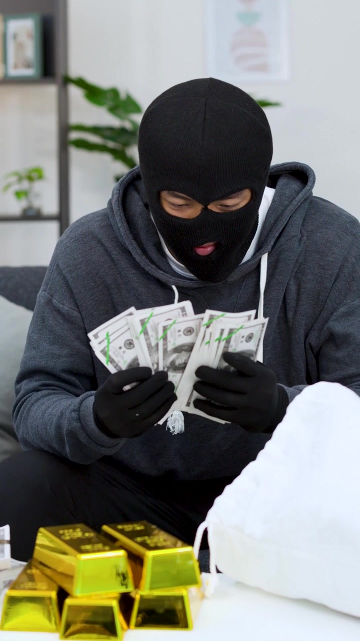 Un ladro con un passamontagna conta banconote da cento dollari accanto a lingotti d'oro con luce intensa.