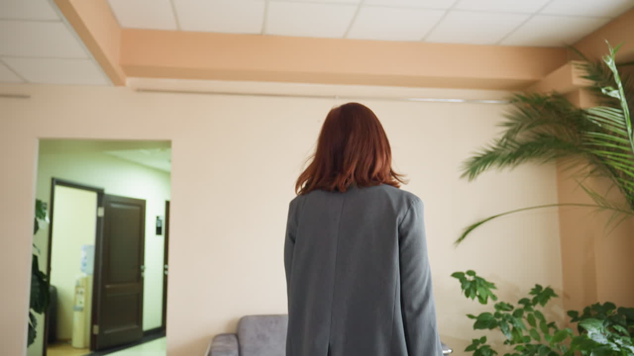 Businesswoman with red hair seen from behind walking toward office hallway entrance in corporate building. Gray suit-clad with cream walls, potted plants, palm decor
