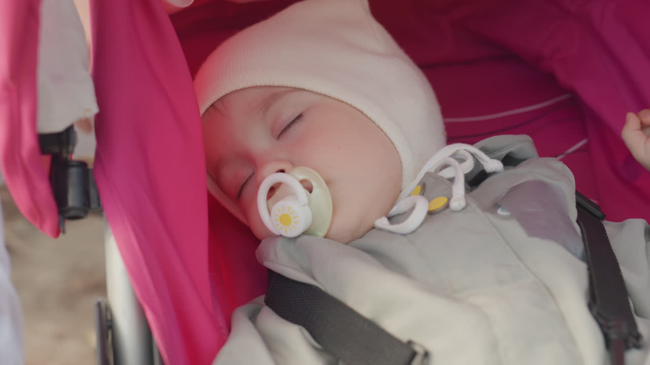 Sleeping newborn in stroller, pacifier visible soft beanie and cozy blanket under pink canopy, peaceful park atmosphere, gentle stroller ride evoking family warmth and parental care