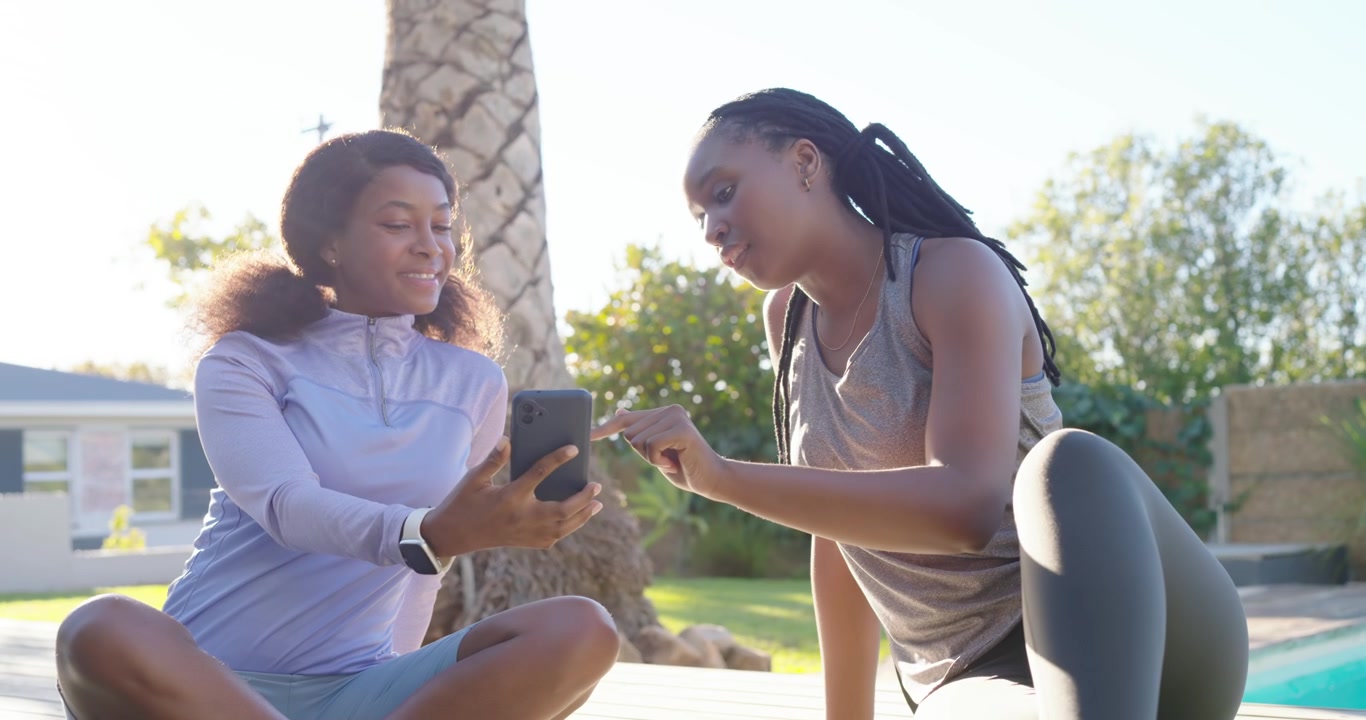 African American women in gymwear sitting deck sharing smartphone one showing screen other tapping