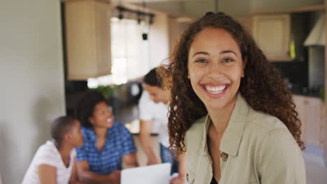 Feliz-Mujer-Birracial-Parada-En-La-Cocina-Y-Mirando-A-La-Cámara-Con-Diversos-Amigos-En-Segundo-Plano.