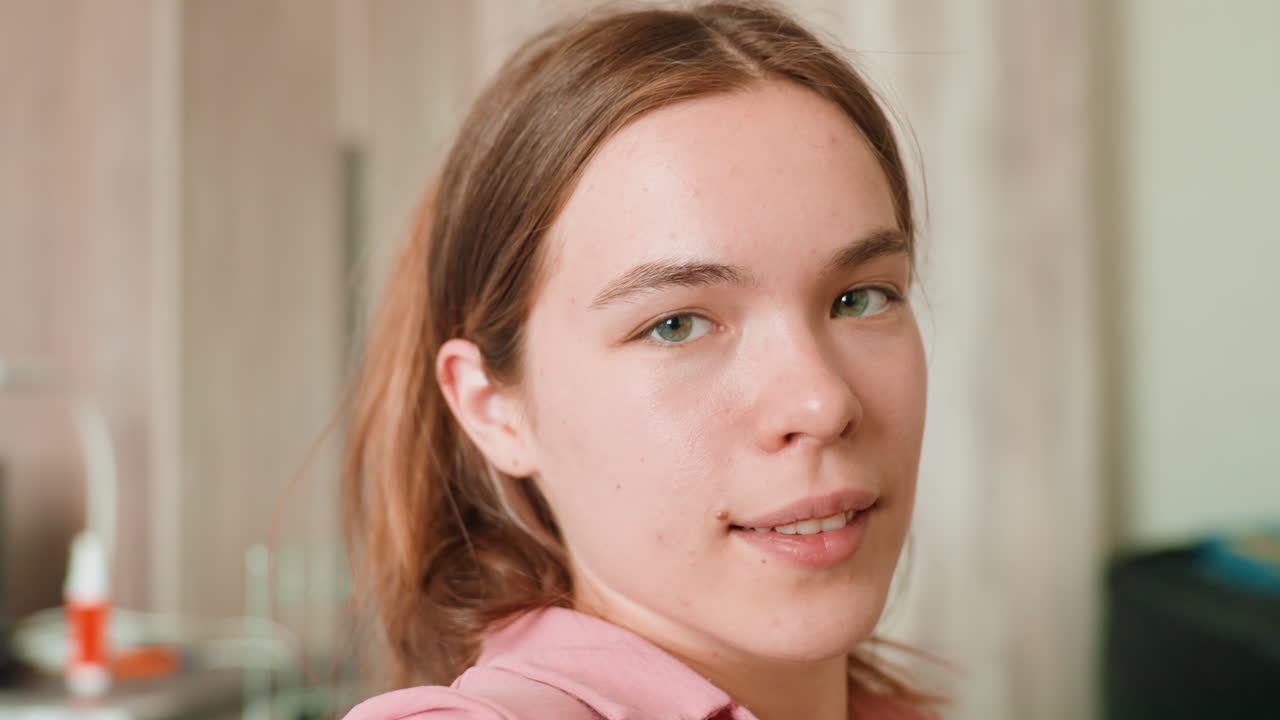 Caucasian Young Woman Adjusting Hair In Livingroom, Pink Hoodie, Natural Light On Face, Soft Background Blur, Introspective Gaze, Relaxed Domestic Mood, Closeup On Expression And Skin
