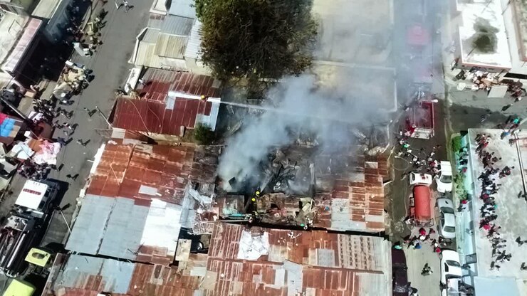 Vista aérea con dron de humo gris elevándose, mientras los bomberos extinguen un incendio estructural humeante en una ciudad; toma de dron circular.
