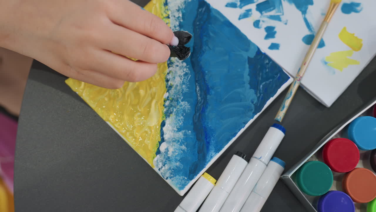 손이 해안선을 그리다, 여성의 손이 작은 캔버스에 파란색 물감을 톡톡 찍어 해안선의 디테일을 만들고, 여성의 손이 어두운 파란색 물감을 스펀지로 덧발라 해안선 질감을 묘사하는 모습 클로즈업