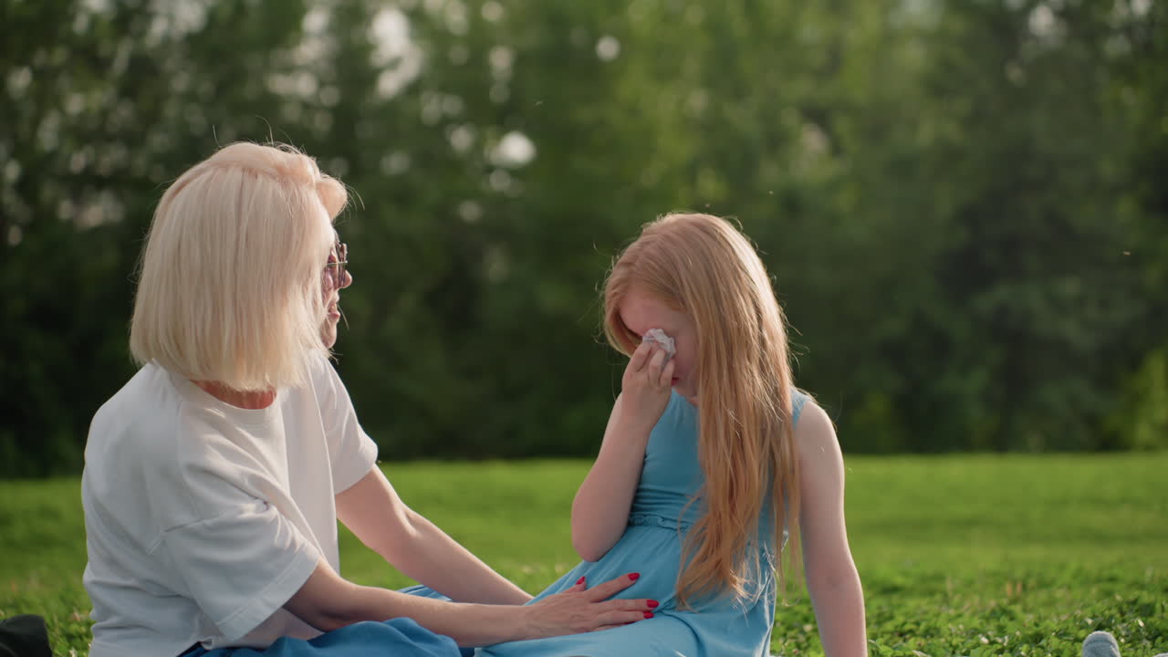 summer girl wiping tears with tissue while mom comforts and cheers her up on grass, soft light and blurred green background creating tender moment of support and resilience