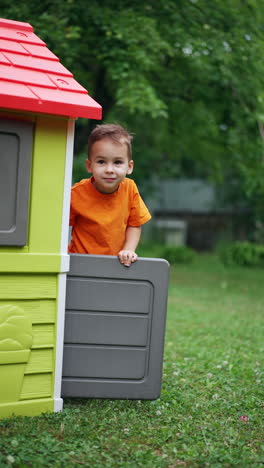 little baby boy leaves the plastic playhouse outdoors. toddler kid looks at camera standing at the doors. vertical video.
