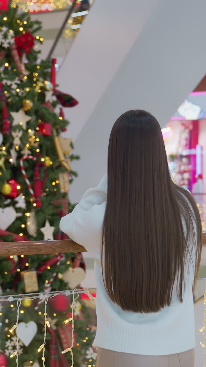 Rear view of woman resting on railing adorned with festive lights, gazing at beautifully decorated Christmas tree in shopping mall, retail ambiance with warm holiday atmosphere and people walking by