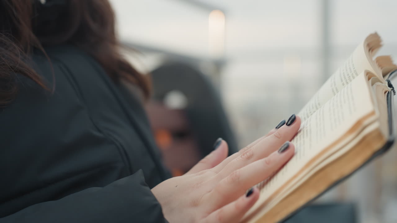 Primo piano mani che leggono un libro usurato, morbidi movimenti delle dita che sfogliano pagine ingiallite, smalto nero, cappotto scuro, sfondo sfocato dello skyline cittadino, ritmo cinematografico lento, atmosfera intima e riflessiva, tattile.