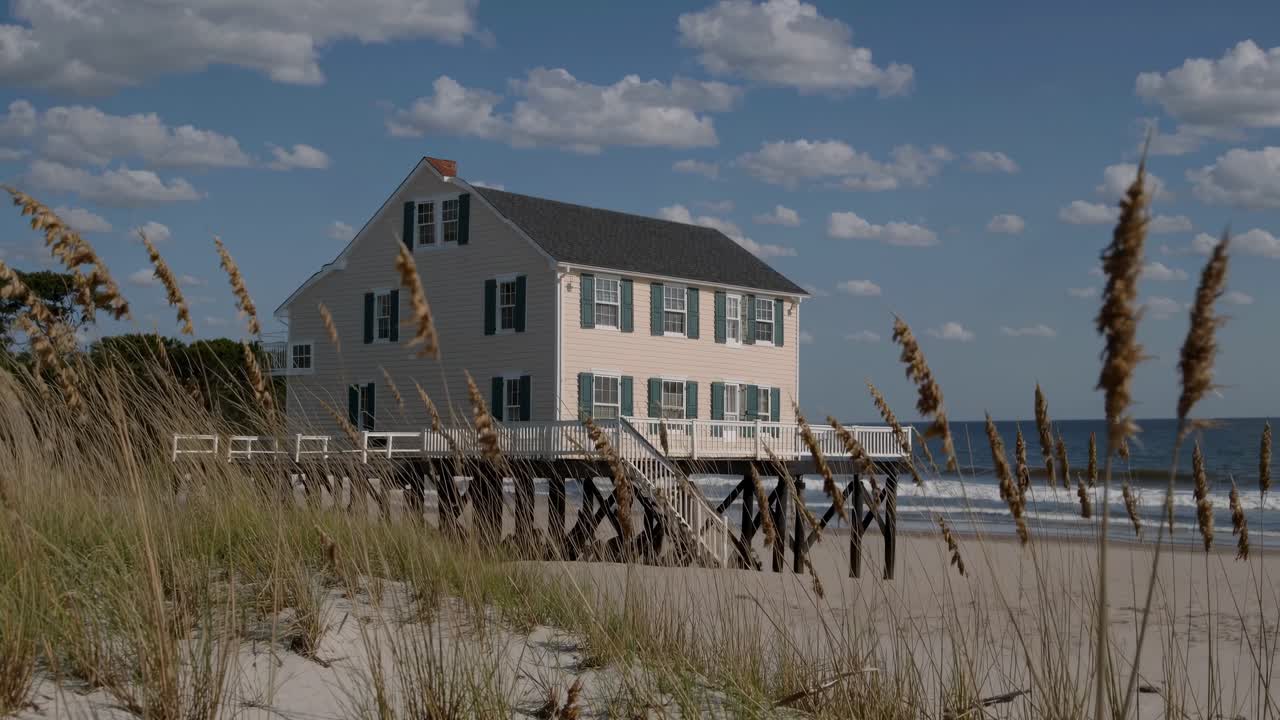 Beach house perched on stilts, offering stunning views of the ocean, with gentle waves rolling in under a sunny sky filled with fluffy clouds and beach grass swaying nearby