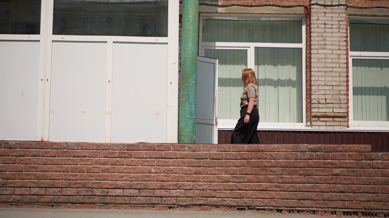 Side view of pupil walking into building carrying handbag under bright sunlight, brick wall and wide windows reflecting peaceful academic mood and everyday