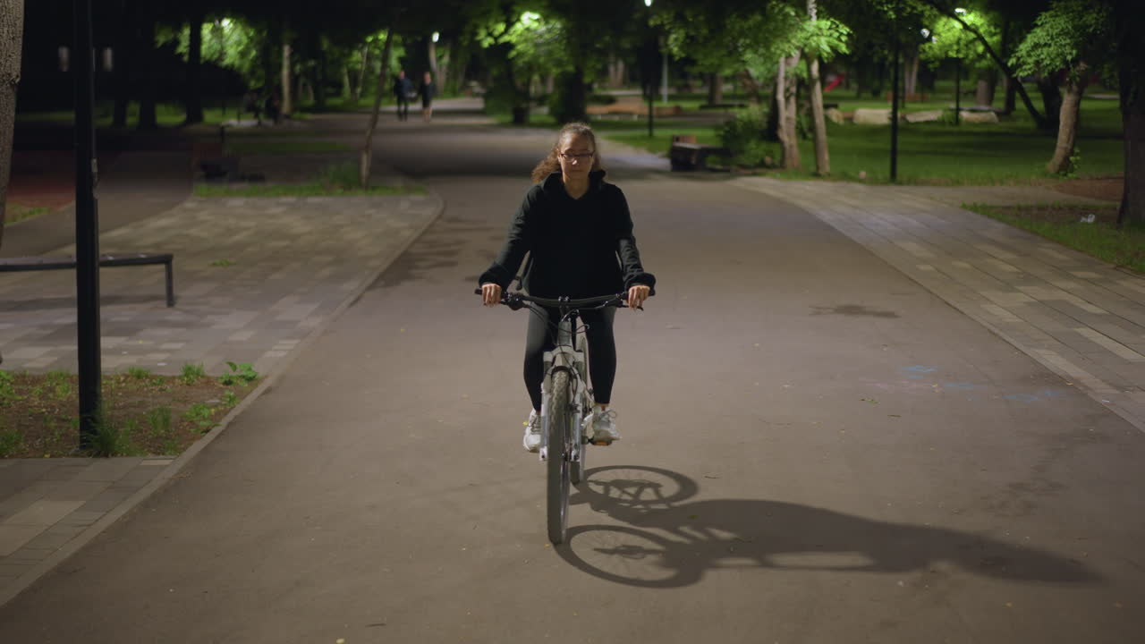 Cyclist Strides Confidently, Woman Rides Along Illuminated Pathway, Confident Female Cyclist On City Walkway At Dusk, Woman Pedals Purposefully Through Lit Urban Promenade At Night