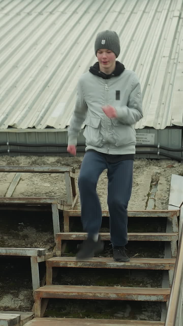 A young boy wearing a gray sweater and a black beanie works out by running up an old rusty iron staircase, the background features a building across the road and a car passing