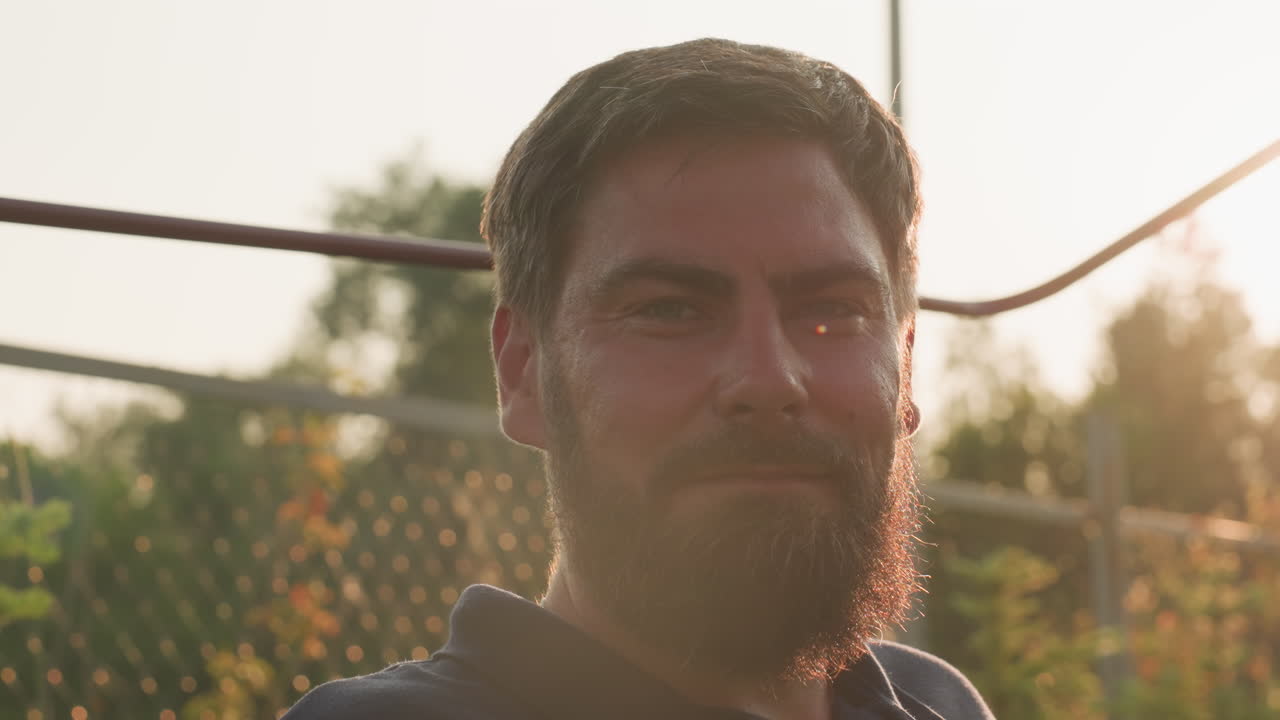 close up of man sitting quietly on swing bench in garden surrounded by trees and fence during golden hour, sunlight creating warm glow around his beard and shoulders in serene outdoor moment
