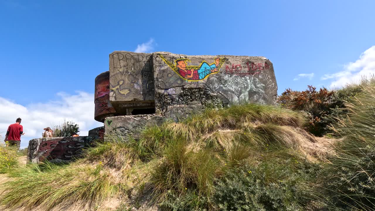 Camera glides through grassy dunes to graffiti-covered concrete bunker as person in red enters