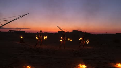 a group of men and woman fire show at night on the sand against the background of fire and tower cranes