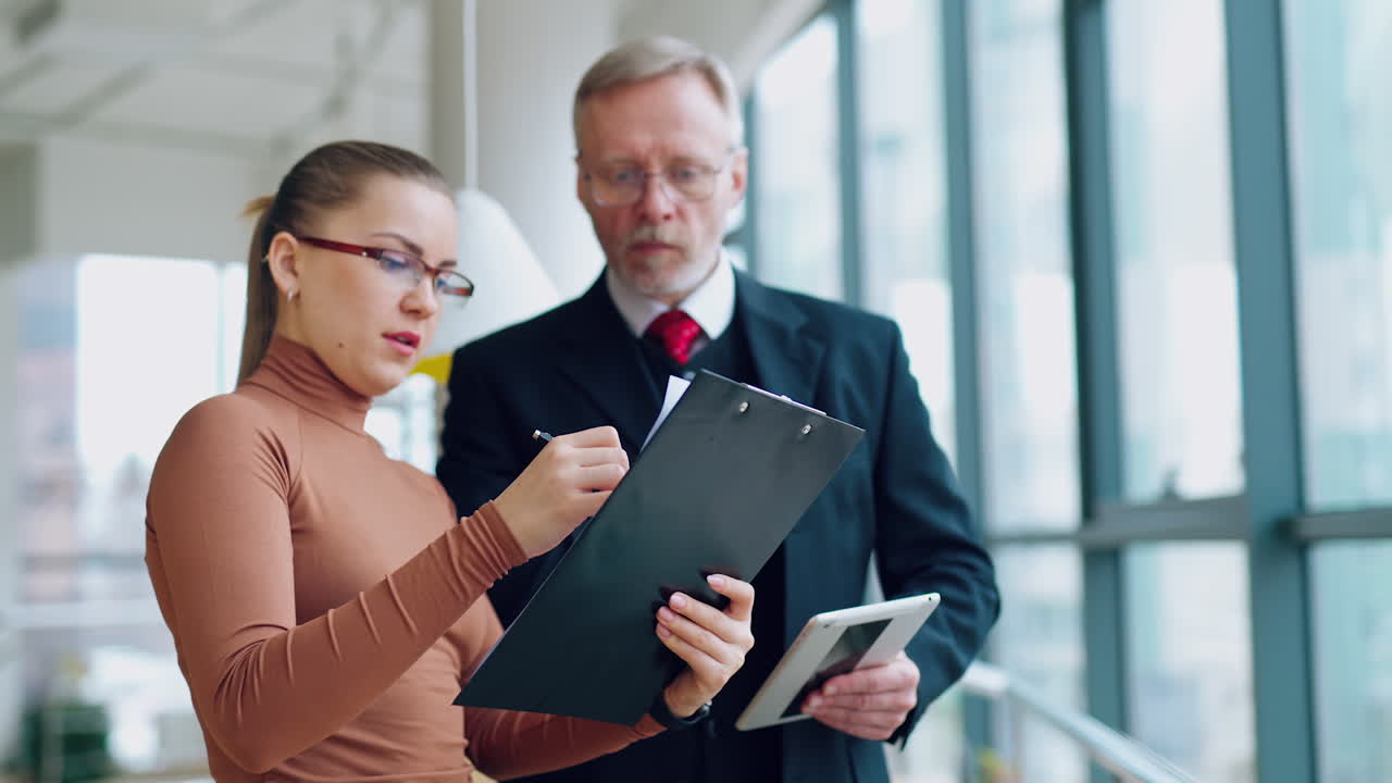Mature man in suit with young secretary indoors. Attractive woman holding the folder and telling her chief about the new project.