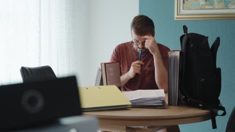 a man sits at a desk, holding a pen to his lips, thoughtfully reviewing paperwork—focused on solving a problem or making an important decision