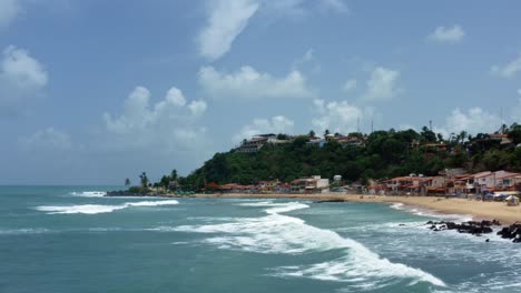 Dolly-in-aerial-drone-wide-landscape-shot-of-the-tropical-famous-Baia-Formosa-beach-town-in-the-state-of-Rio-Grande-do-Norte,-Brazil-with-fishing-boats,-coastal-homes,-small-waves,-and-surfers