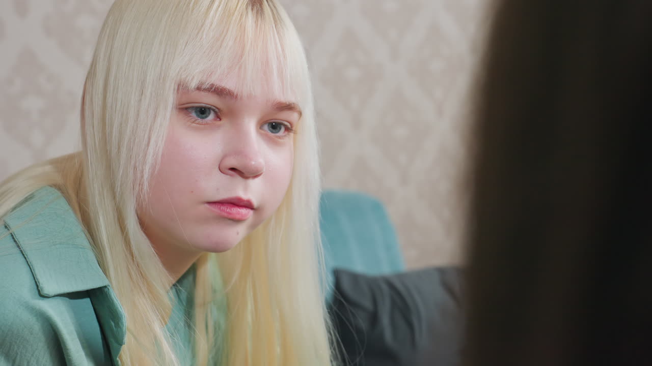 Close up head view of young woman with blonde hair wearing green shirt eating popcorn and talking to person near her on patterned background in indoor casual setting, natural light scene