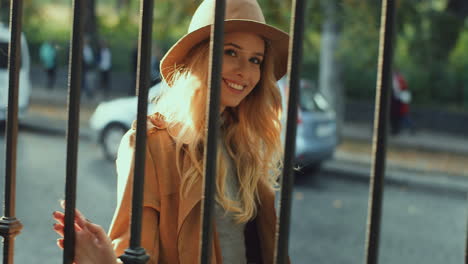 cheerful smiled blonde young woman weating a hat and coat walking behind a fence in the street