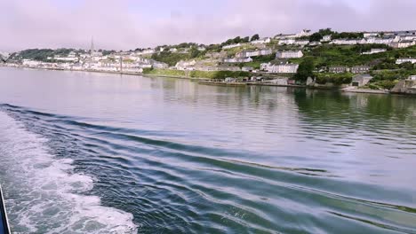 보트로 코브 (cobh) 에 접근하여 아일랜드 코크 카운티 (cork county) 에 있는 마을 위의 파노라마와 함께 파도를 만들고 있습니다.