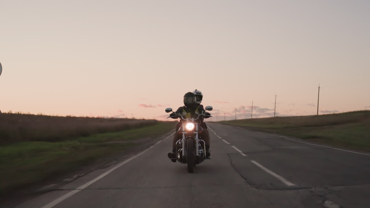 Two bikers ride motorcycle straight toward camera on open countryside road during colorful sunset, adventure, and travel with glowing headlight and wide rural horizon
