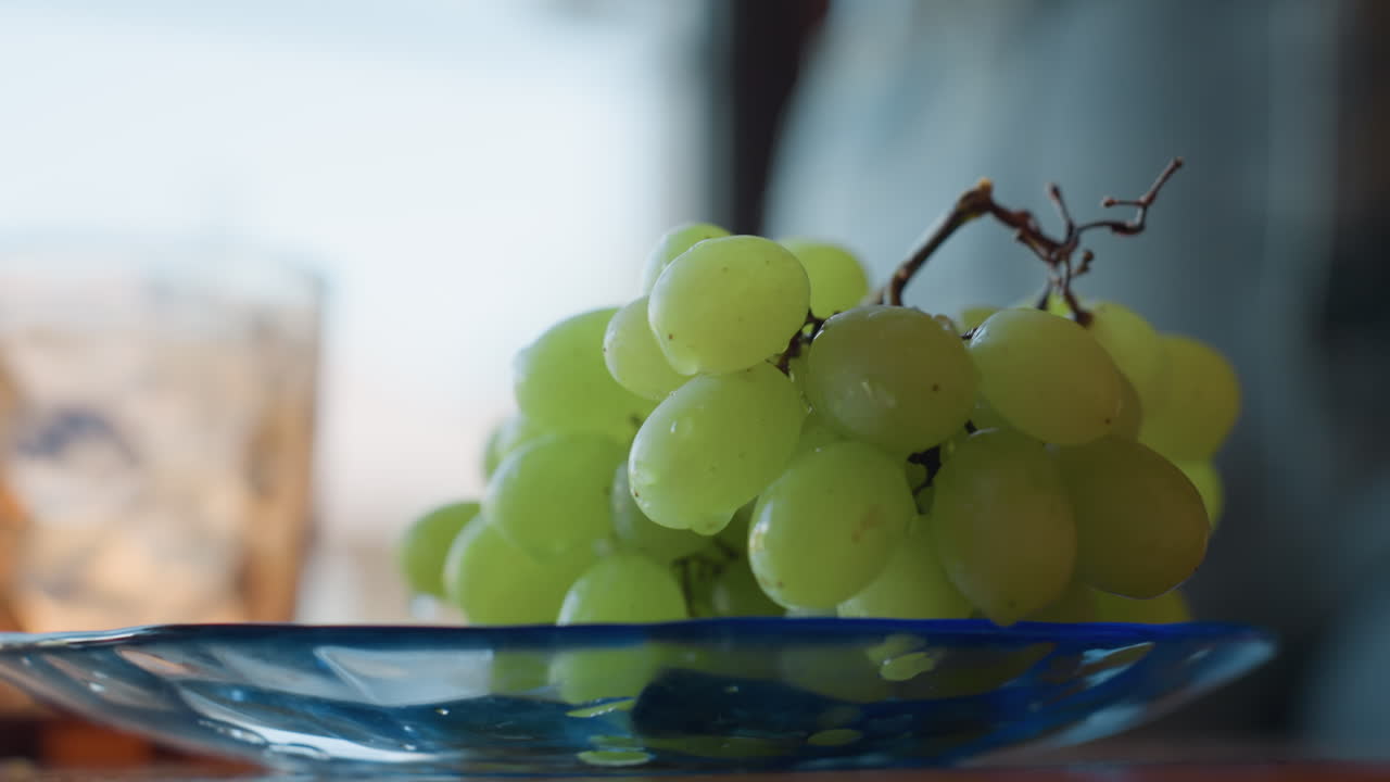 Close up of fresh green grapes being dropped onto transparent blue glass plate set on wooden table, with soft lighting highlighting juicy texture and moisture on fruit
