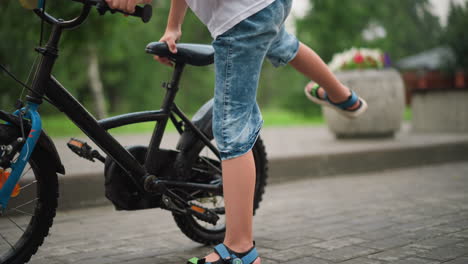 a young boy is seen from the legs down, as he approaches and climbs onto a bicycle, wearing denim shorts and colorful sandals, the child rides off with the bicycle on a paved path