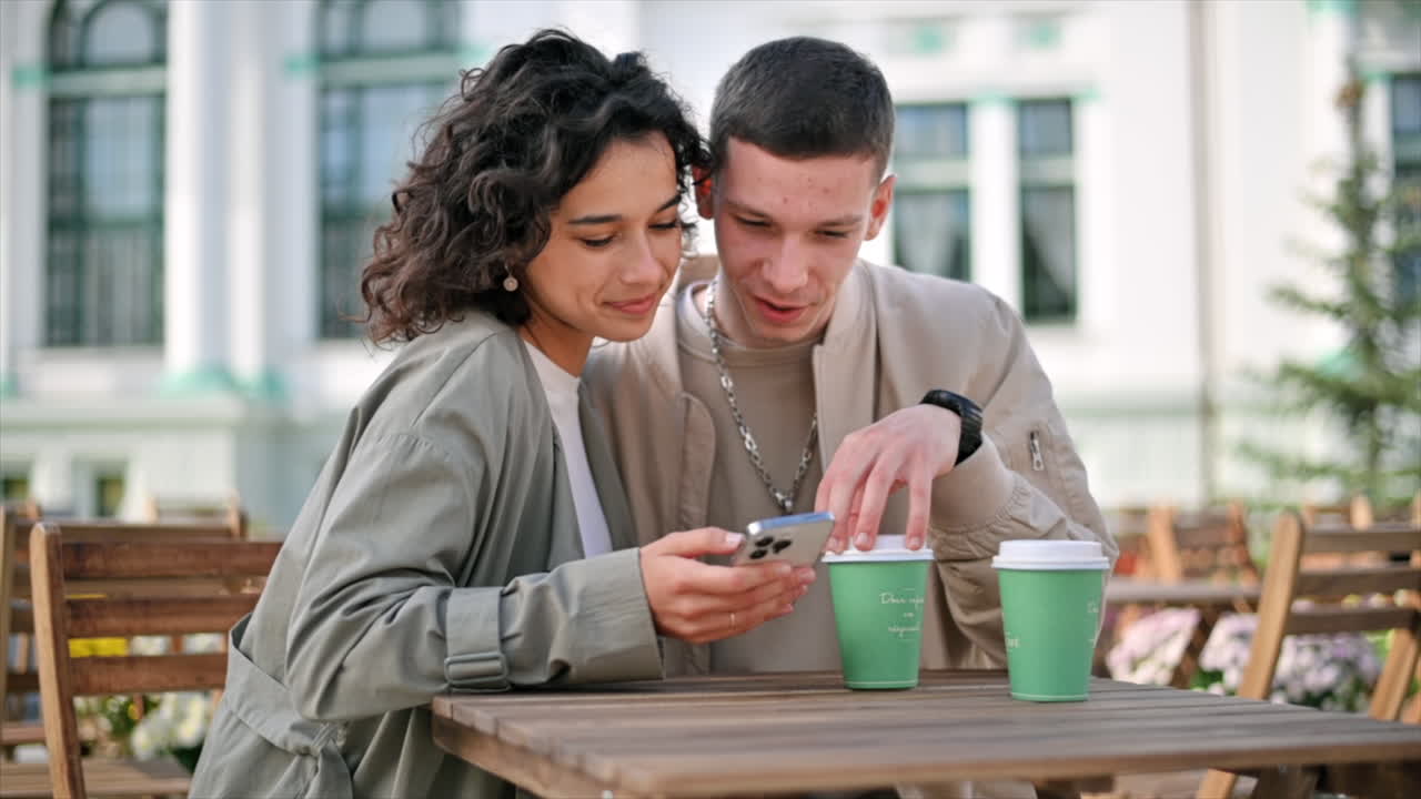 A happy couple outdoors near a cafe. Looking in the phone, smiling, talking, coffee on the table. Autumn atmosphere. Slow motion