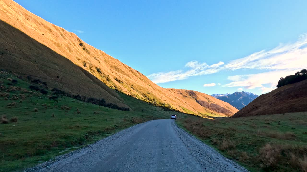 Vehicle travels gravel road between sunlit hills, wide landscape, steady camera, natural daylight