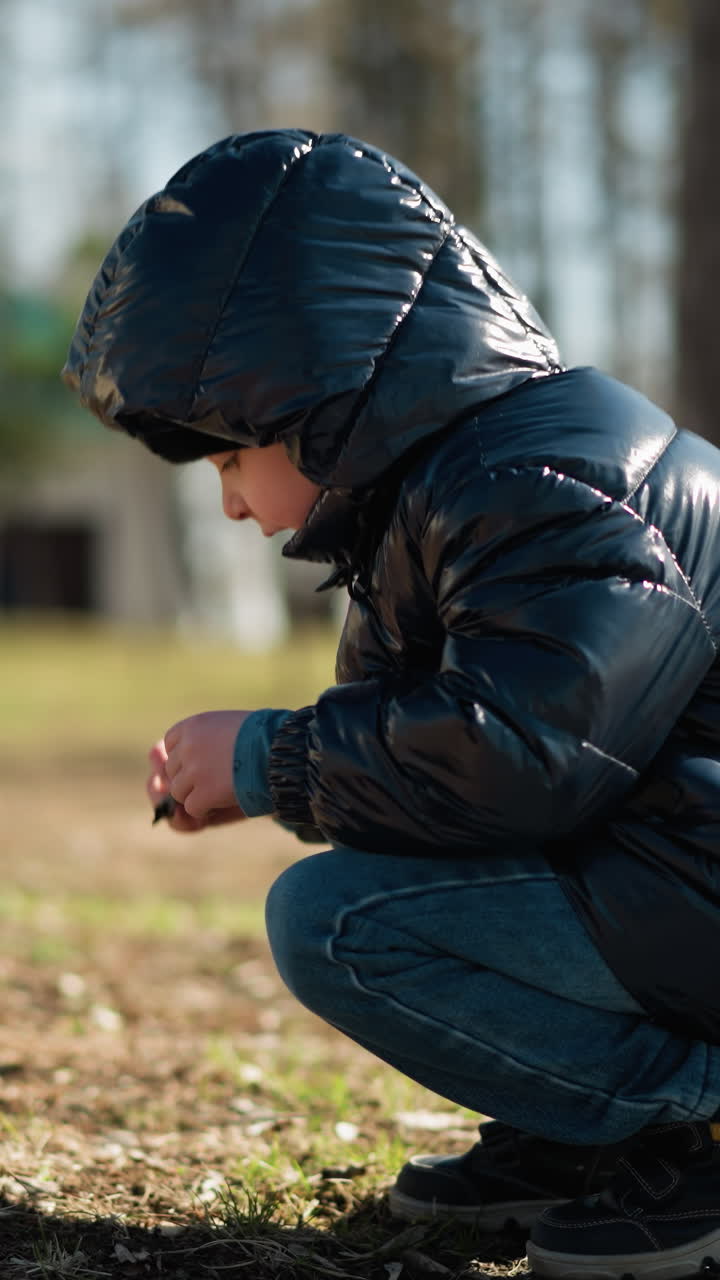공원에서 앉아있는 어린 소년의 회전 시선, 손에 을 집중적으로 관찰하고, 그는 검은색 가죽 재, 헤드 워머, 그리고 청바지를 입고, 나무의 흐릿한 배경으로