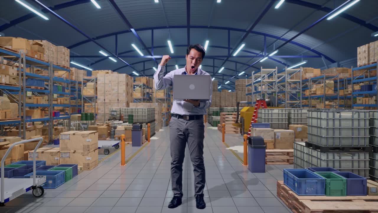 Lavoratore professionista asiatico a figura intera in piedi con il suo laptop in un magazzino con file di scaffalature, alza il pugno in segno di vittoria.