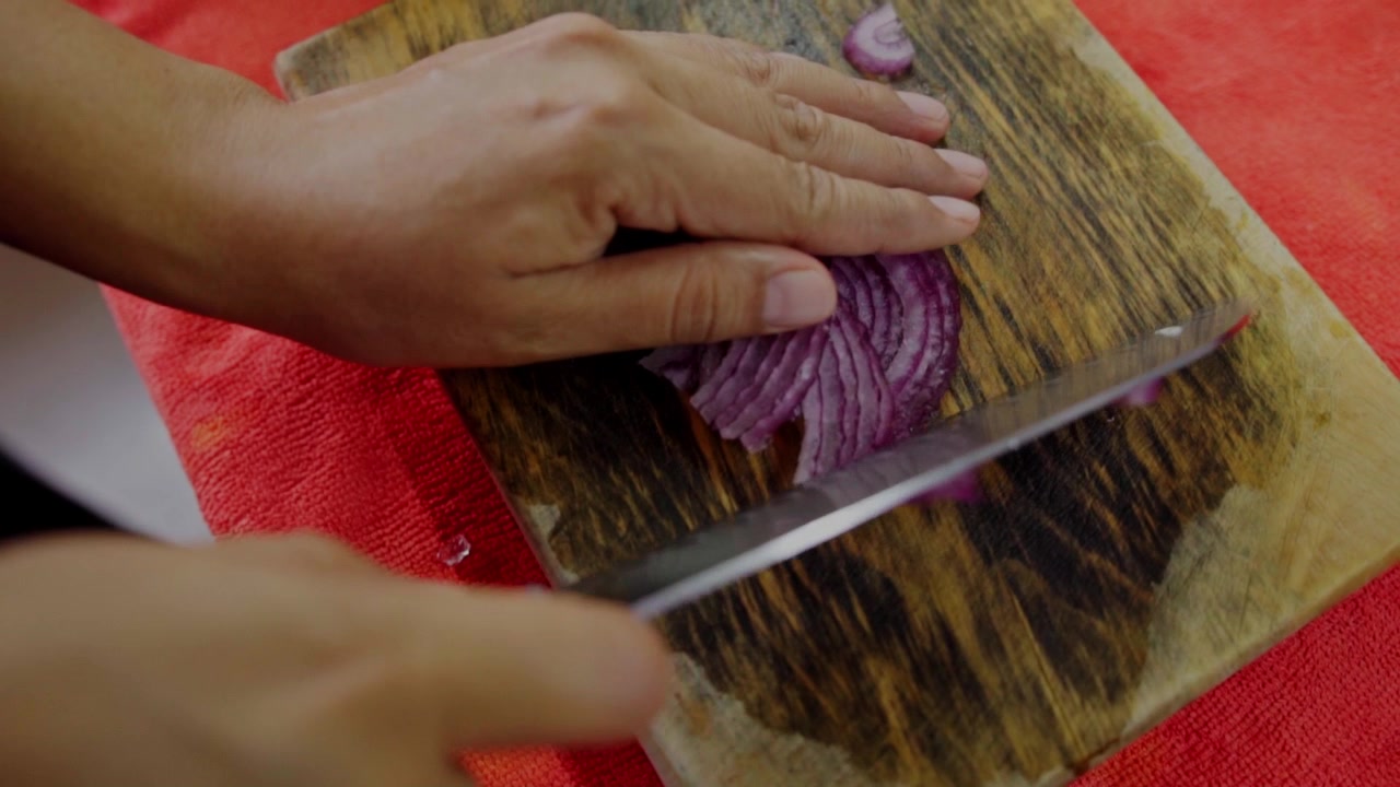 Closeup view of person slicing red onion with knife on wooden cutting board during homemade meal preparation in kitchen