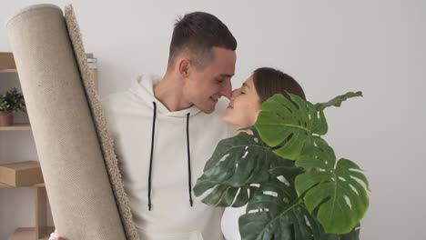 zoom in shot of young affectionate couple in a new house holding a carpet and a plant 1