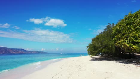 Pristine-beach-with-white-fine-sand-under-shadow-of-lush-vegetation,-washed-gently-by-white-waves-of-blue-sea-on-a-shiny-sky-with-overhanging-clouds-in-Indonesia