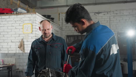 mechanic in workshop working on car engine, tightening nut with red gloves while colleague observes, background features work tools, yellow wall clock, and bright light illuminating workspace