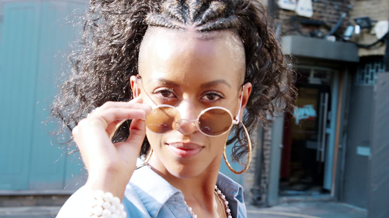 파란 드레스를 입은 트렌디한 젊은 흑인 여성이 그녀의 선글라스를 통해 카메라로 돌아보고, 가까이 다가갑니다.