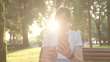 Eine-Frau,-Die-Durch-Ihr-Telefon-Scrollt,-Während-Sie-Im-Park-Sitzt,-Mit-Sonnenblende-Hinter-Ihr,-Handheld-Aufnahme