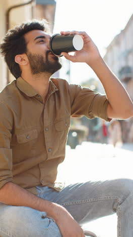 young indian man enjoying drinking morning coffee hot drink relaxing taking a break in city street