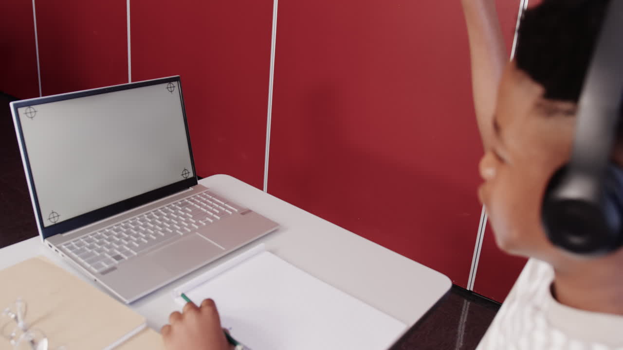 In school, African American teenage boy wearing headphones using laptop and writing in notebook, cop