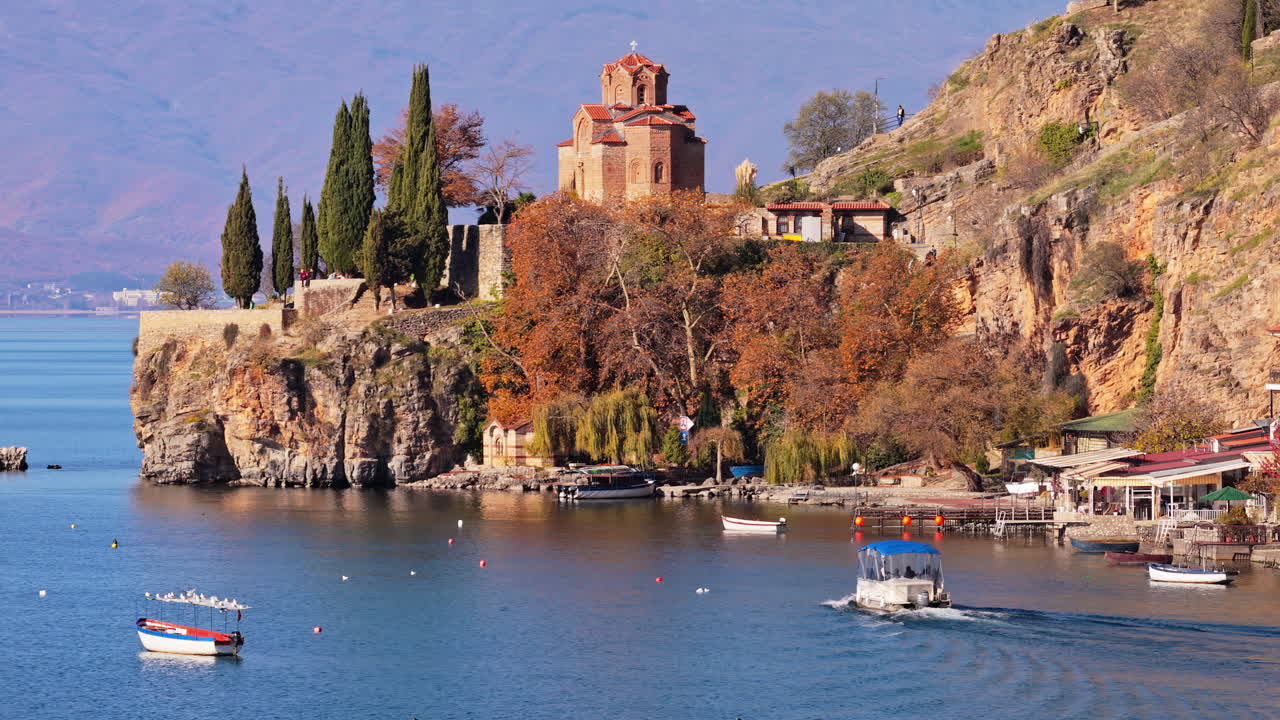 Aerial drone view of the Church of St. John at Kaneo, surrounded by rocky cliffs, autumn trees, and small boats docked along the shoreline
